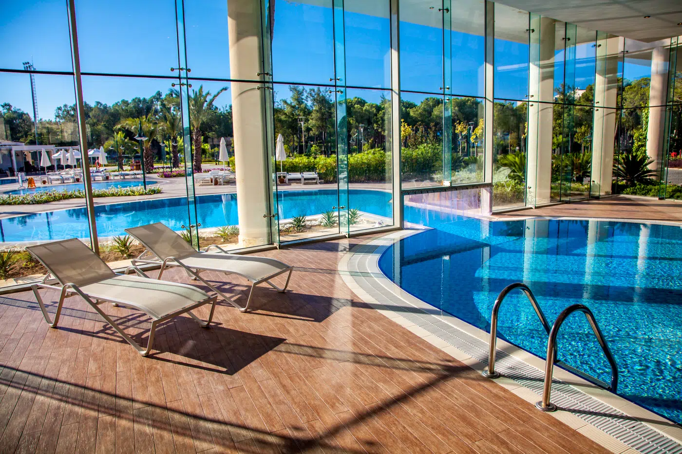 Sunlit glass-walled resort swimming pool pavilion with loungers